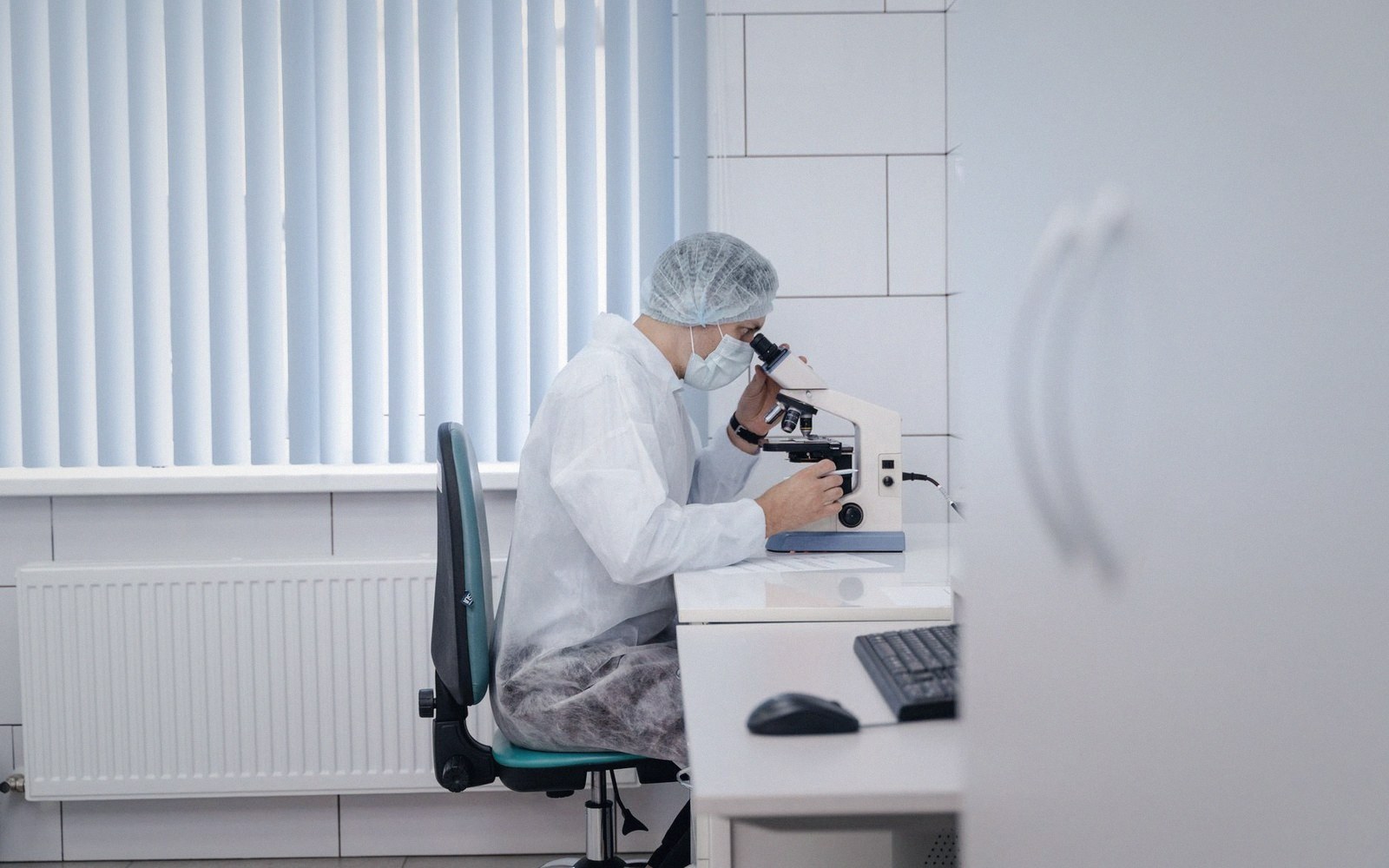 Lab technician in protective gown, mask and hairnet examining a sample under a microscope
